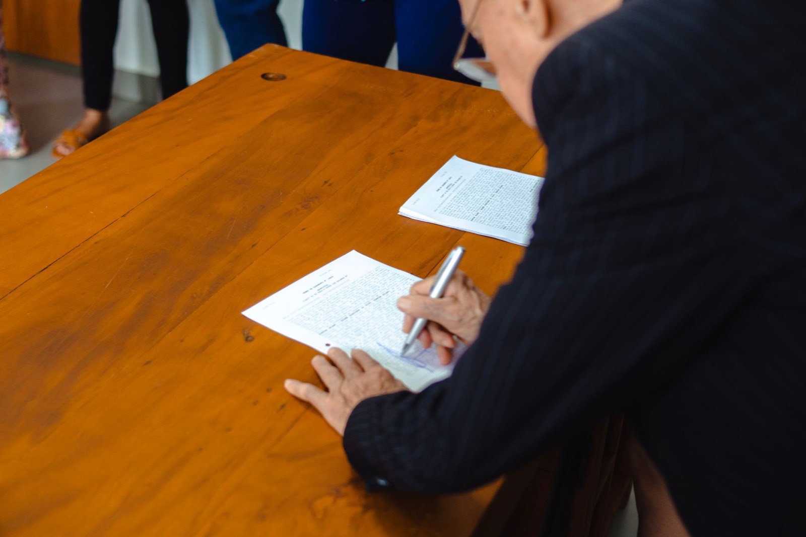 A person in a dark jacket signs printed documents at a wooden desk while colleagues stand nearby
