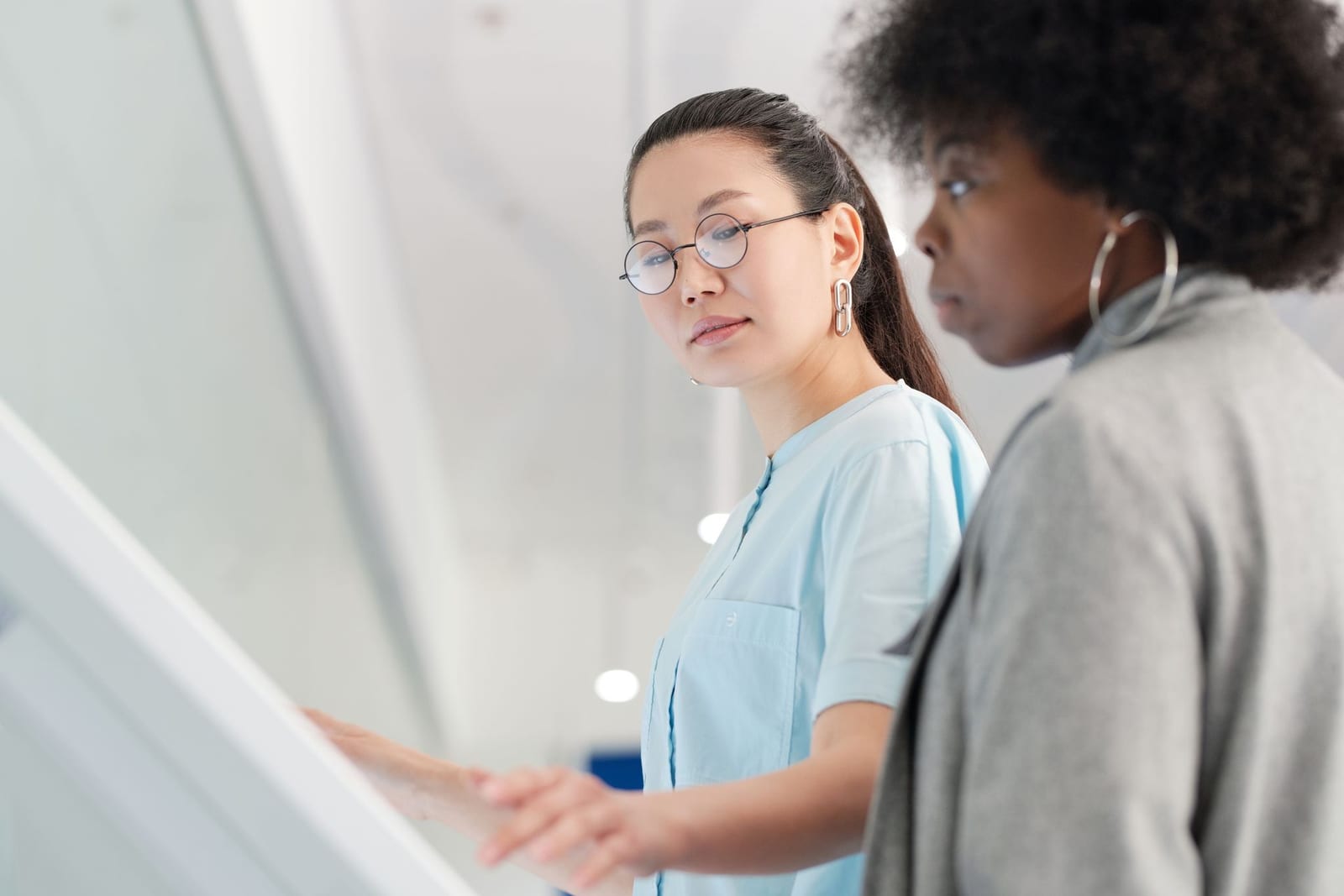 A clinician in a light blue coat points to something on a bright wall-mounted screen while a colleague looks on