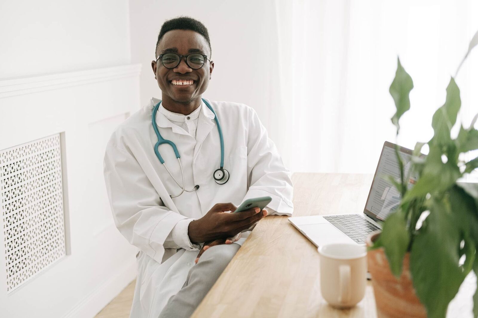 A clinician in a white coat smiling while checking a phone beside an open laptop in a bright office