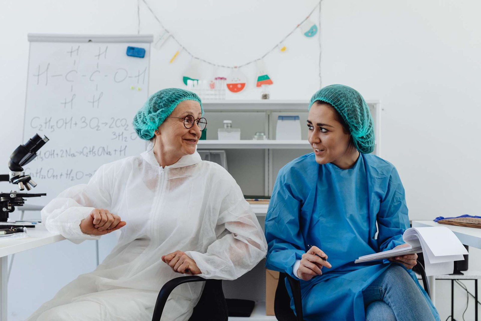 Two researchers in cleanroom hairnets and gowns talk beside a whiteboard covered in chemistry notation