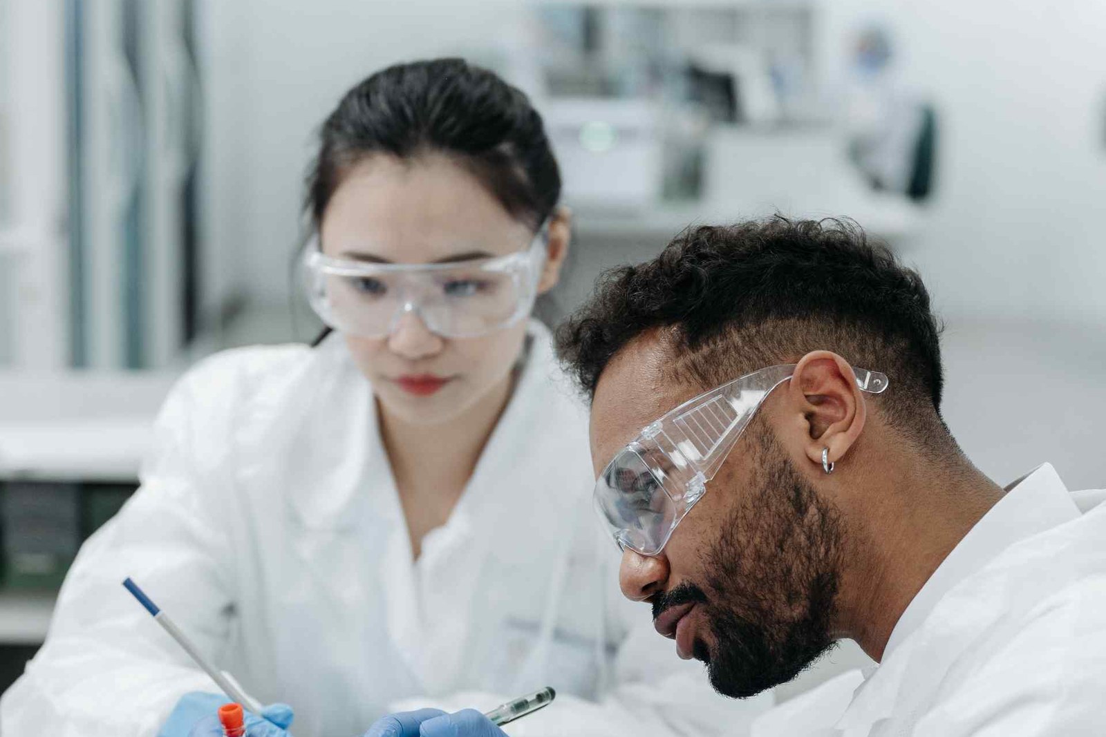 Two lab specialists in white coats and safety glasses examine a blood-collection tube in a clinical laboratory