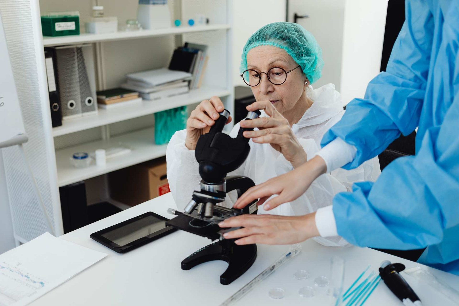 A researcher in a lab coat adjusts a microscope while a colleague in scrubs assists, examining a sample under magnification