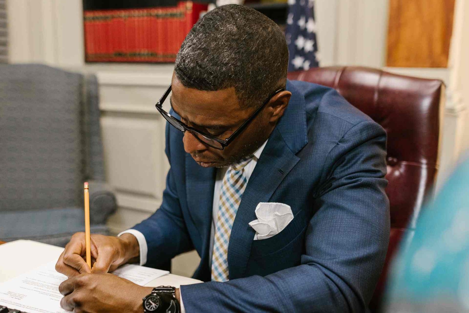 A man in a suit reviews and signs a paper document at a desk, with an American flag in the background