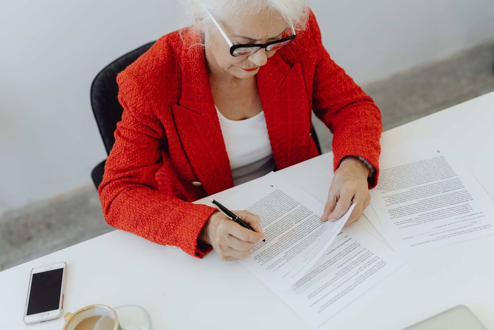 A woman in a red blazer reviews and signs printed documents at a white desk