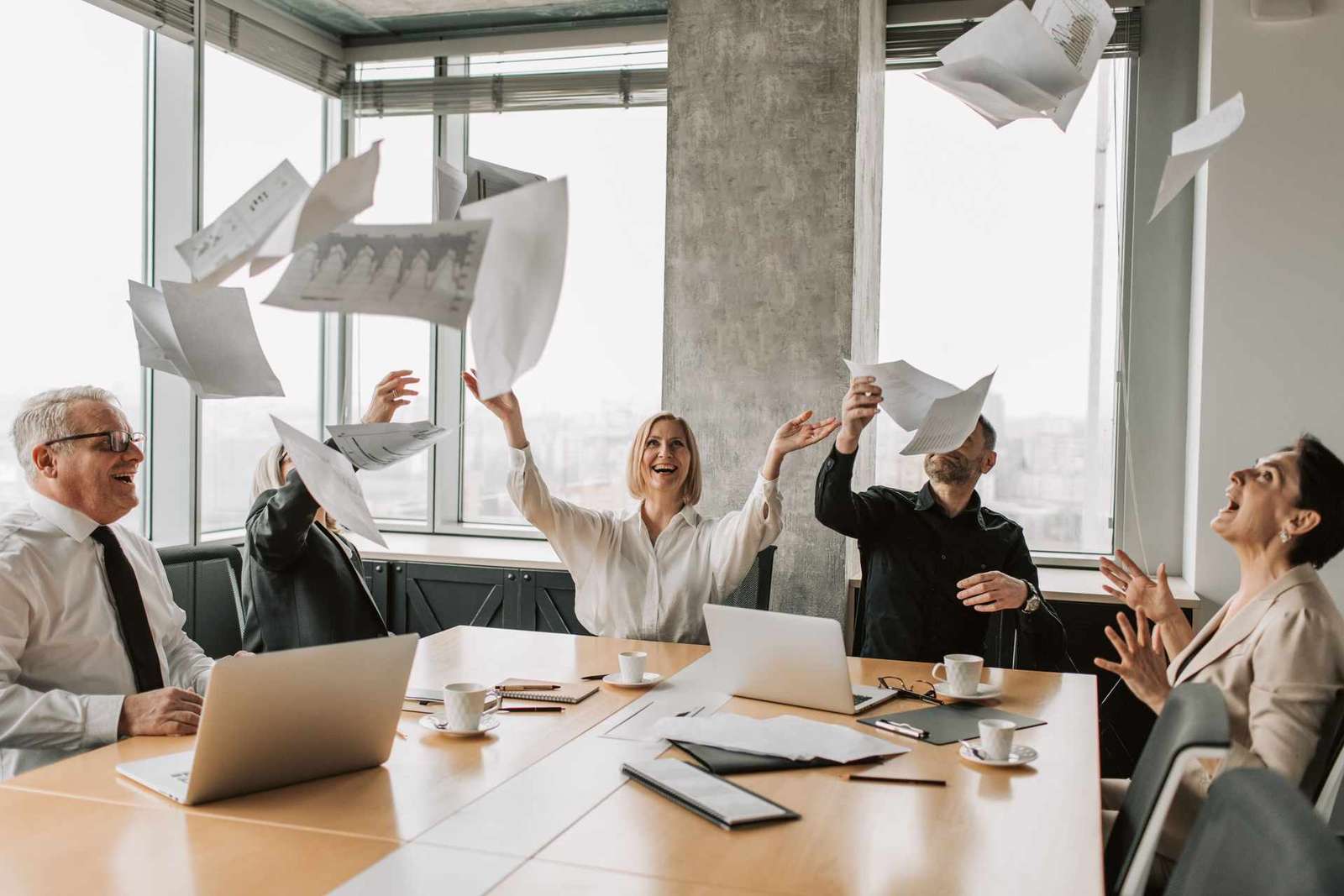 A team of colleagues in a bright office celebrate a milestone, tossing papers into the air around a conference table