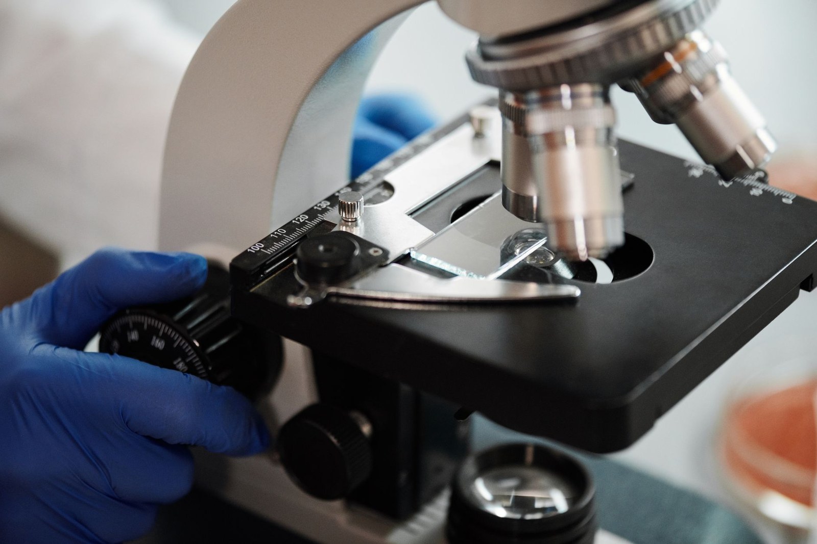 A gloved hand adjusts the focus stage of a laboratory microscope holding a slide