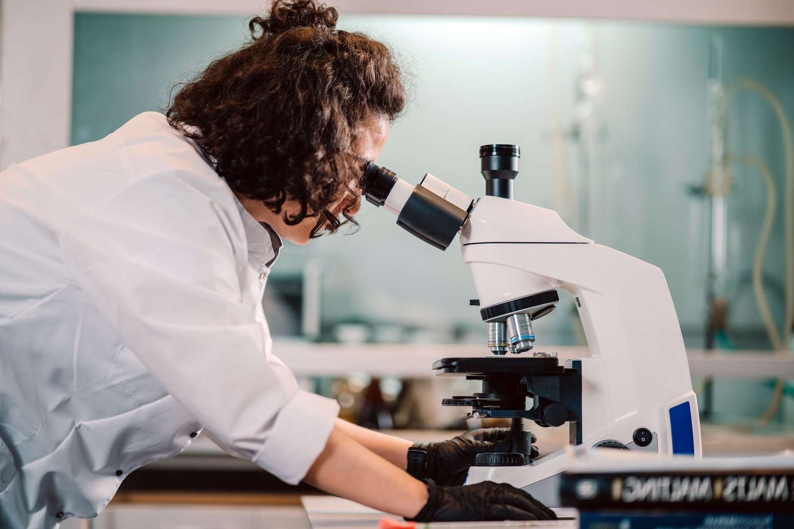 A researcher with curly dark hair in a white lab coat and black gloves looks into a white benchtop microscope in a laboratory