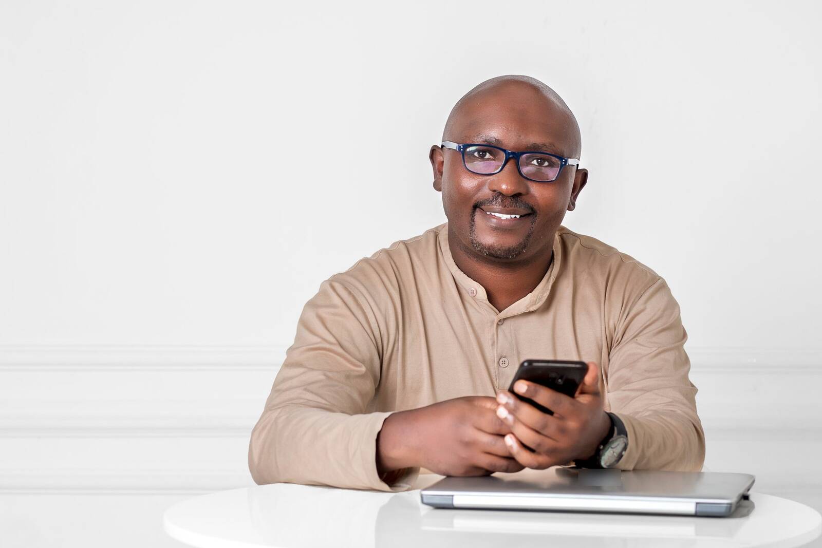 A smiling professional in business attire sits at a white table, glasses on, holding a phone beside a closed laptop