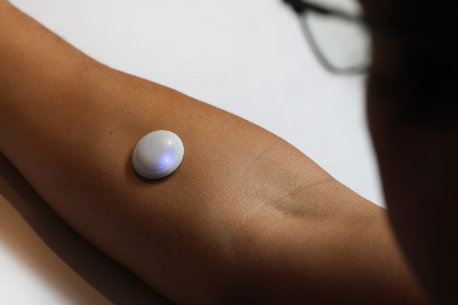 Biolinq Shine, a coin-sized white patch, worn on the underside of a forearm with its LED indicator lit blue