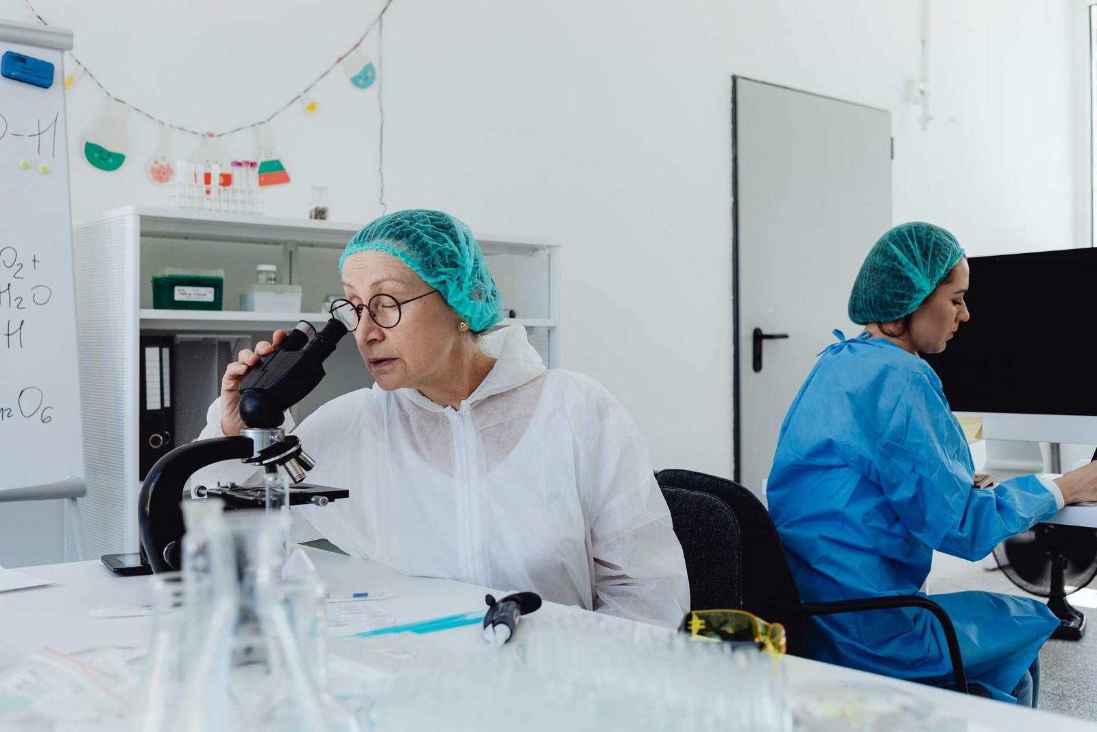 A researcher in a lab coat and hairnet looks into a microscope at a bench lined with glassware, with a colleague working at a computer behind her