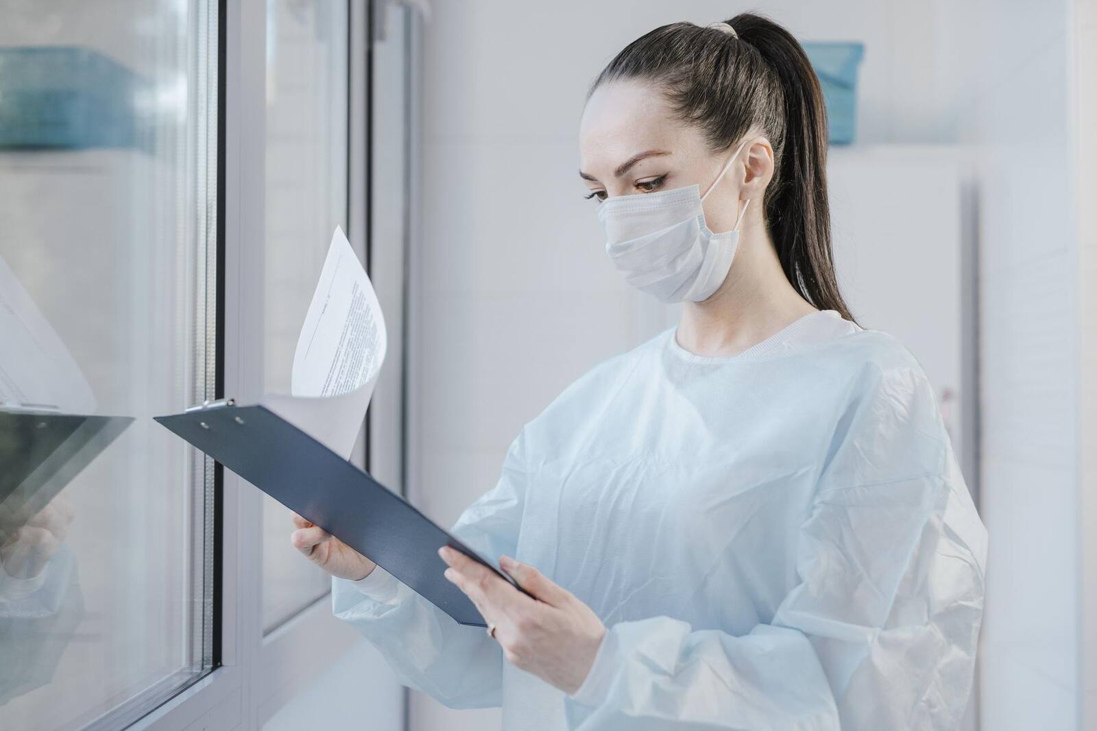 A gowned, masked clinician reviews a chart on a clipboard in a clinical setting