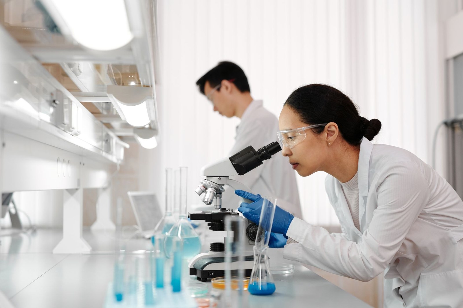 Two researchers in a laboratory, one examining a sample under a microscope while a colleague works at a fume hood in the background