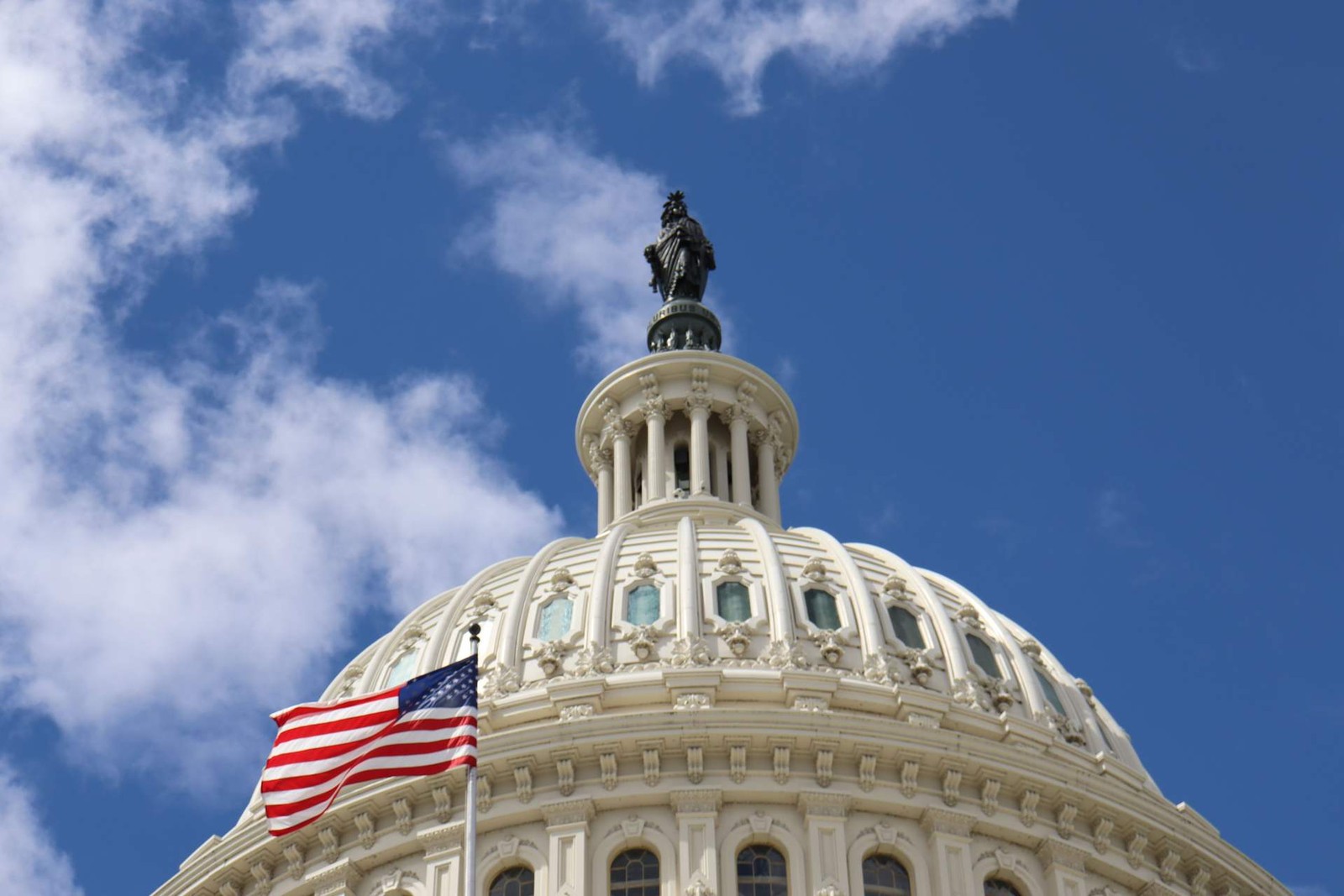 The dome of the U.S. Capitol building with an American flag flying in front against a blue sky