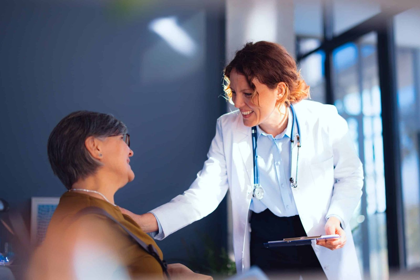 A smiling clinician in a white coat rests a hand on the shoulder of a seated older woman during a consultation, from Celcuity's website