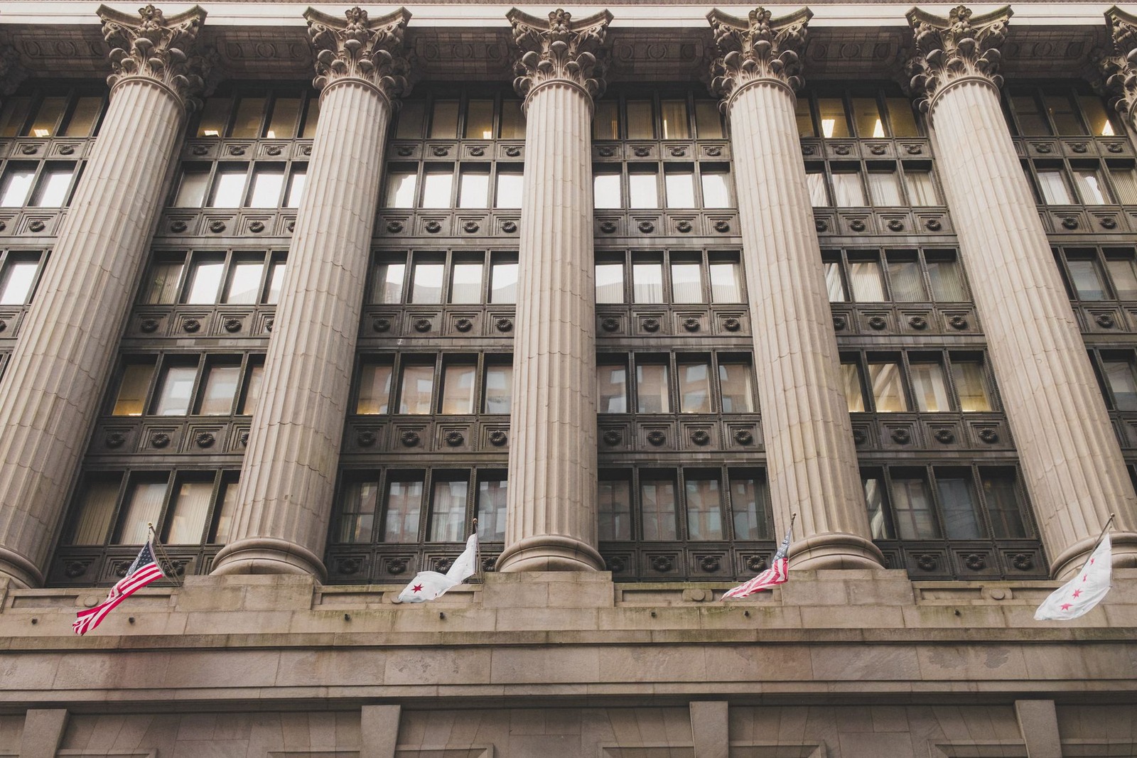 A row of Corinthian columns rises along the stone facade of a government office building flying American flags