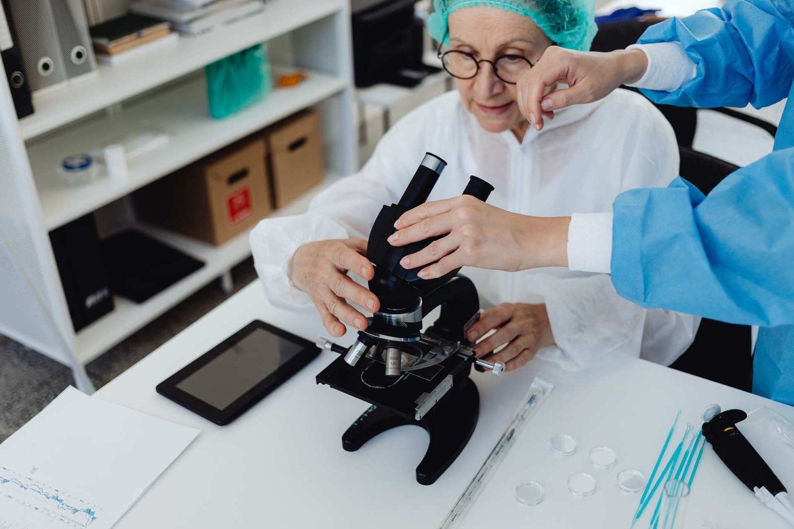 A senior scientist in a cleanroom gown and hairnet adjusts a microscope while a gowned colleague assists at the bench