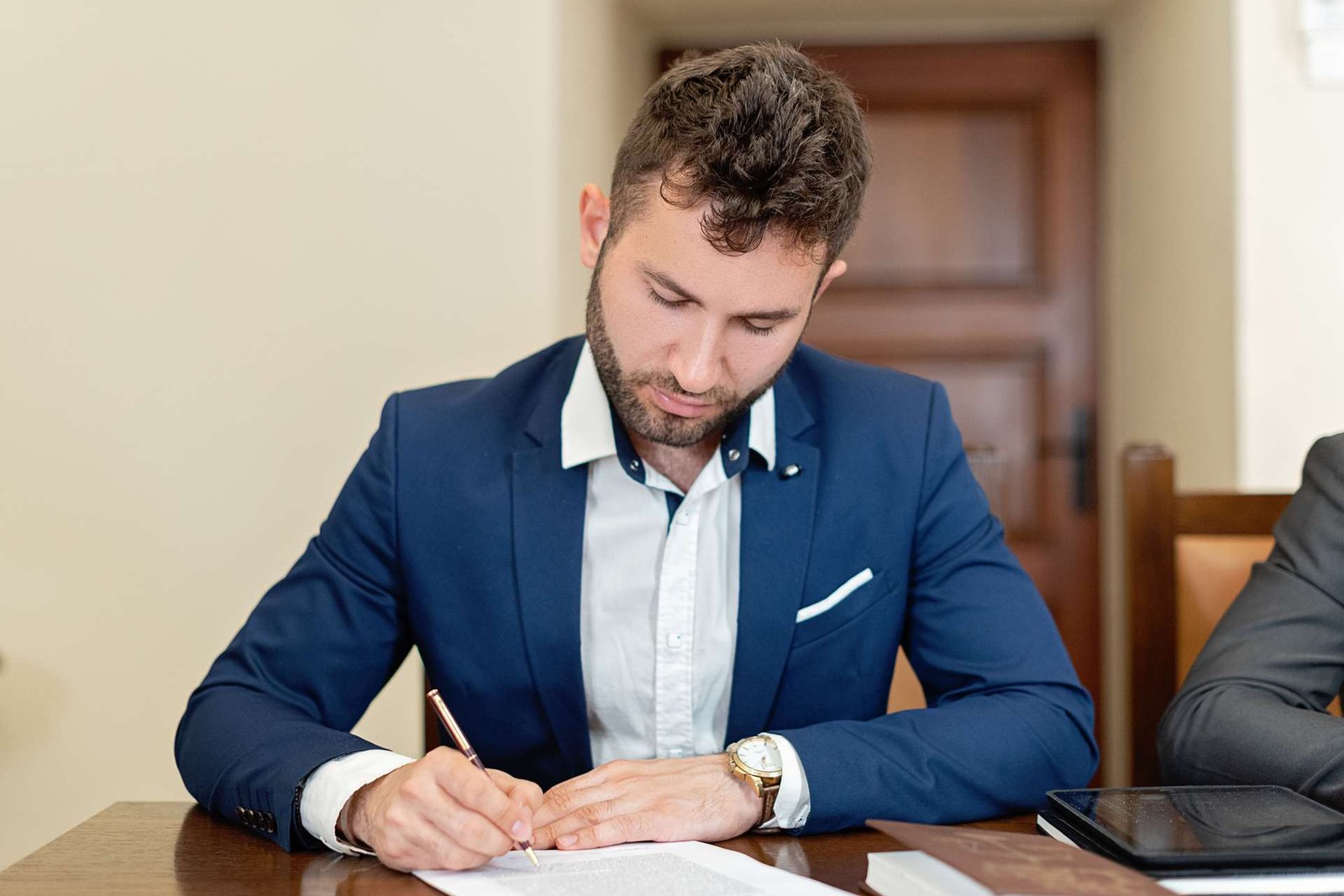 An executive in a navy suit signs an official document at a wooden desk