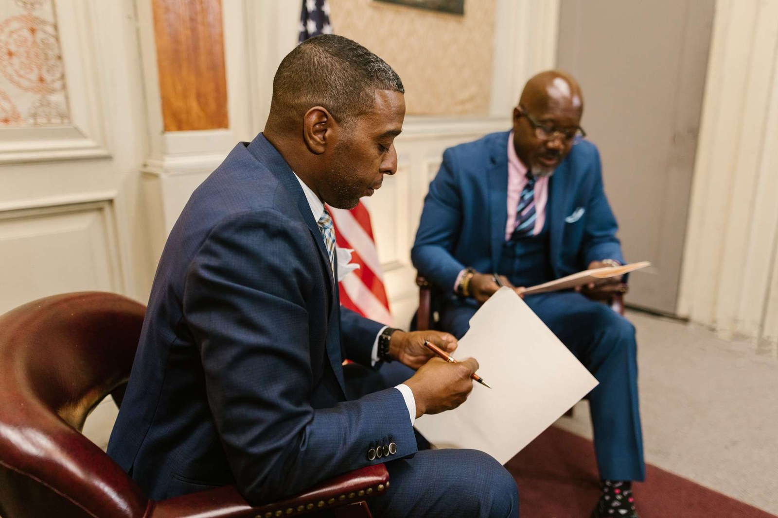 Two men in suits seated with an American flag behind them, one writing on a document held on a clipboard while the other reviews papers