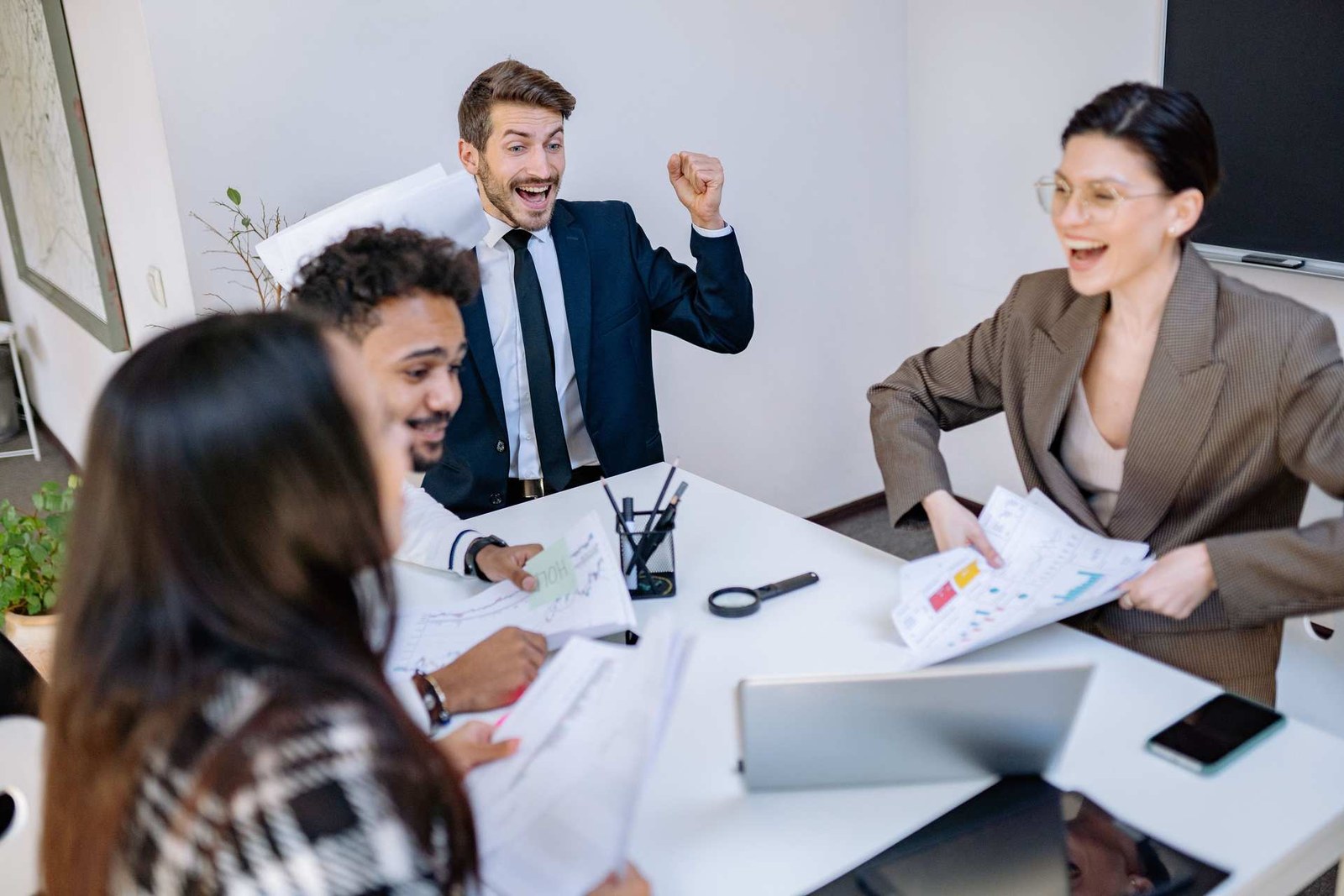 A team of colleagues in business attire celebrate around a conference table with charts and papers