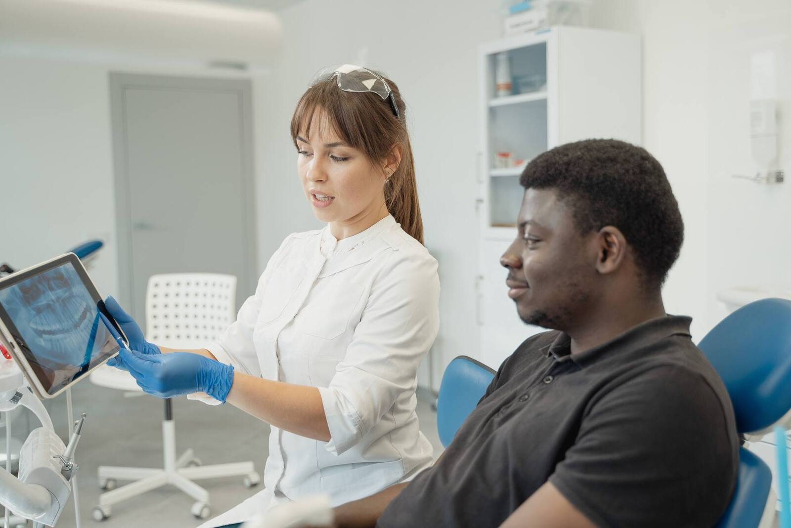 A clinician in a white coat points out findings on a tablet screen to a patient seated in an exam chair