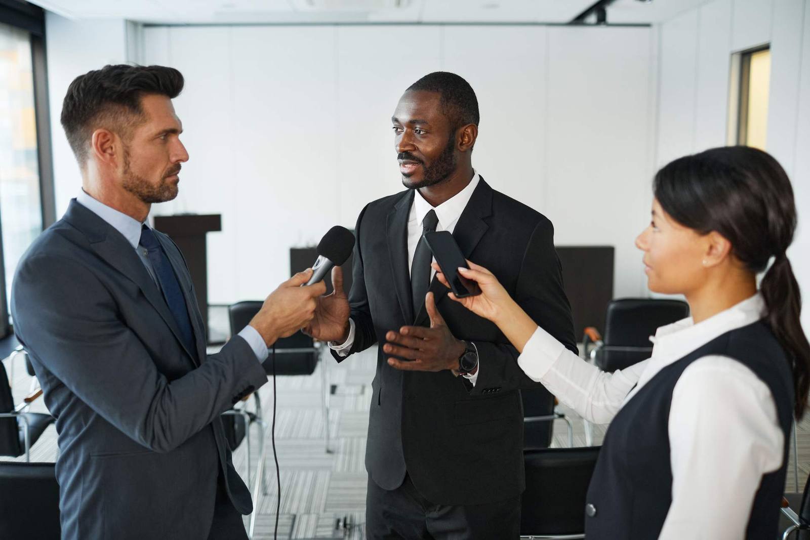 Three people in business attire speak into microphones and a phone recorder held out by reporters in a conference room