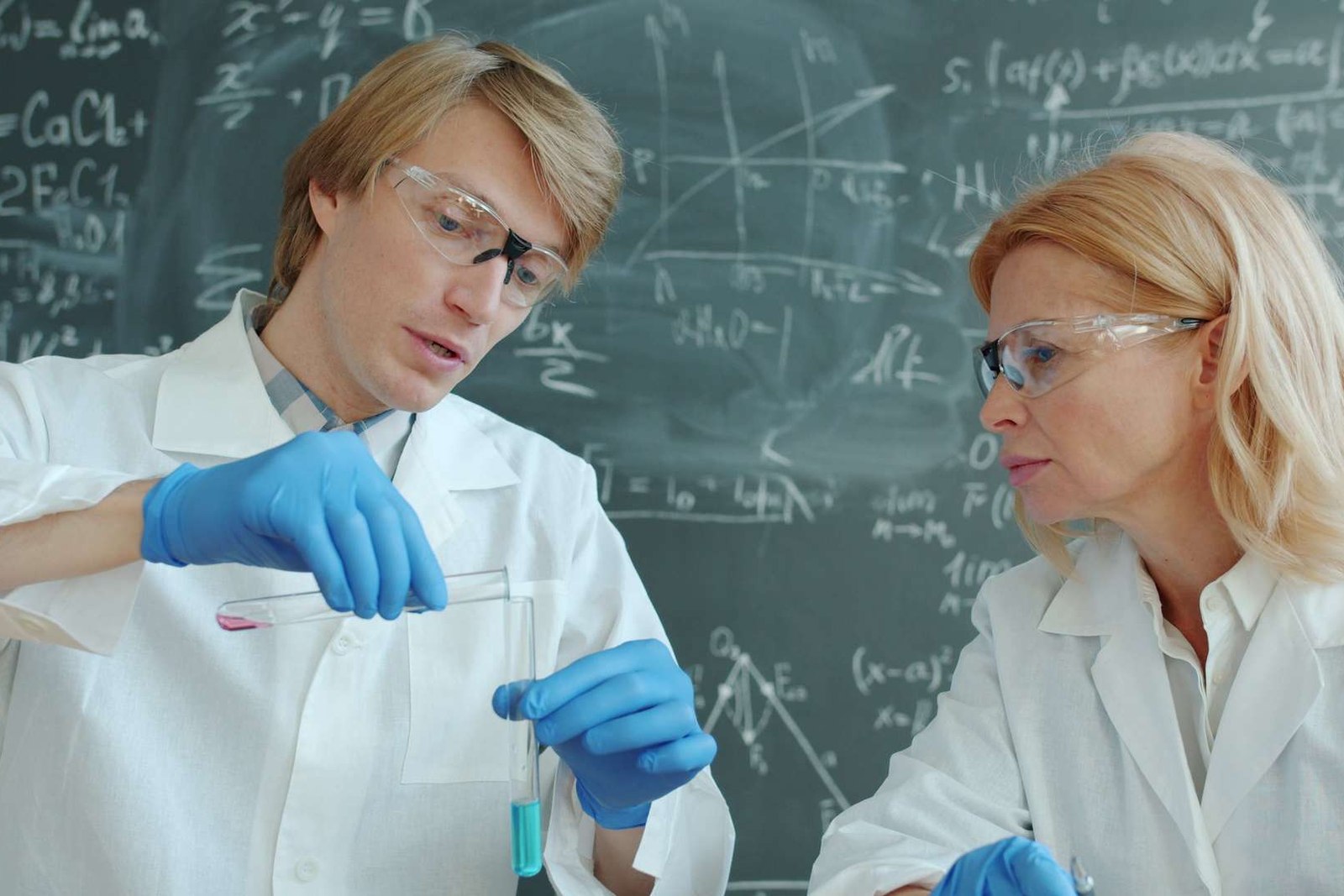 Two scientists in lab coats and safety goggles examine a test tube of colored liquid in front of a chalkboard covered in chemistry equations