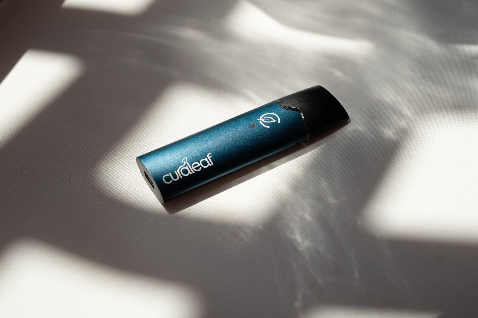 Curaleaf International's QMID inhaler, a slim blue-green metallic device with a dark mouthpiece and the Curaleaf leaf mark, lying on white fabric in dappled sunlight