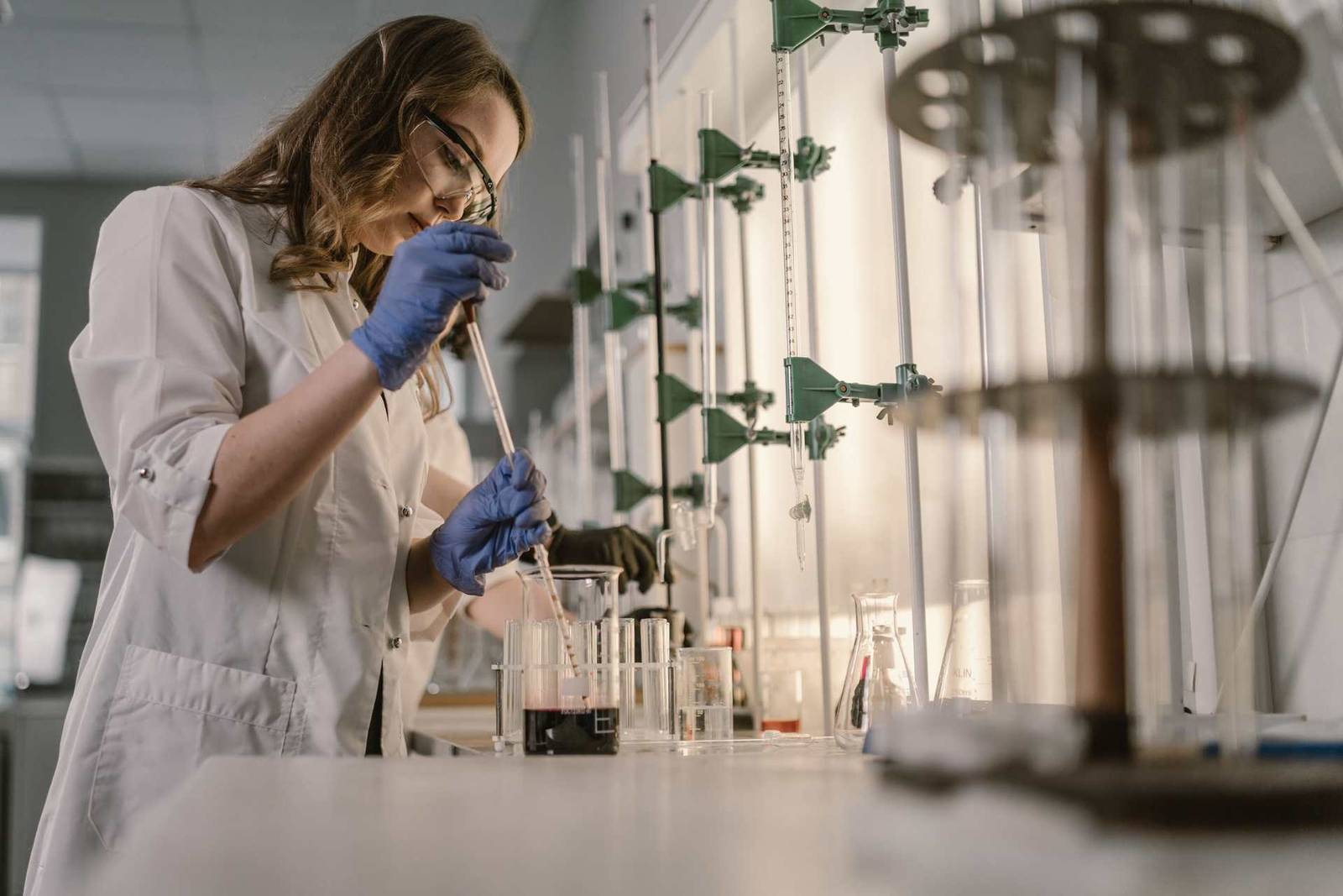 A researcher in a white lab coat and safety glasses uses a pipette over glass beakers and flasks on a laboratory bench