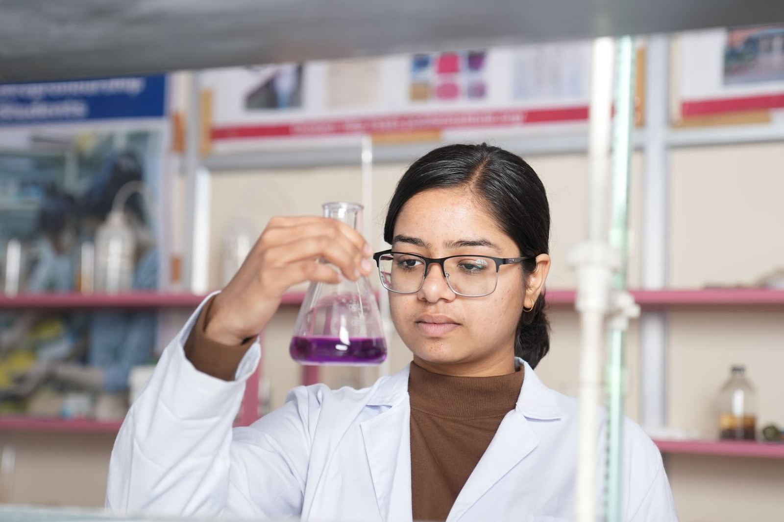 A researcher in a lab coat and glasses examines a flask of purple liquid held up to the light