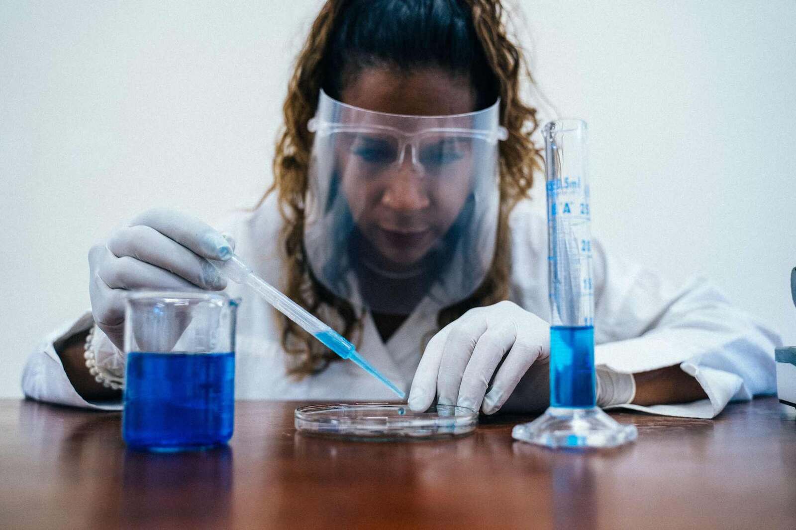 A researcher in a lab coat and face shield uses a pipette with blue reagent, standing in for small-molecule drug research