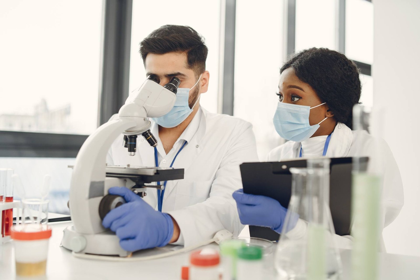 Two masked lab researchers review notes on a clipboard while operating a microscope, surrounded by lab glassware