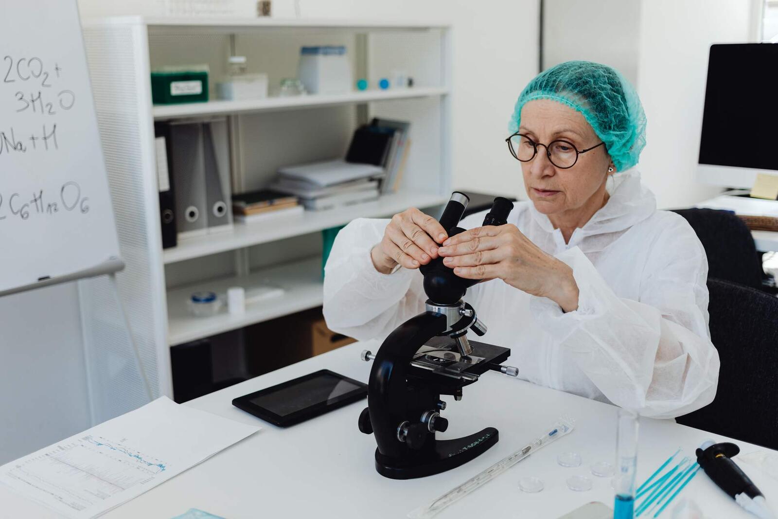 A researcher in a hairnet and lab coat adjusts a microscope at a bright laboratory bench