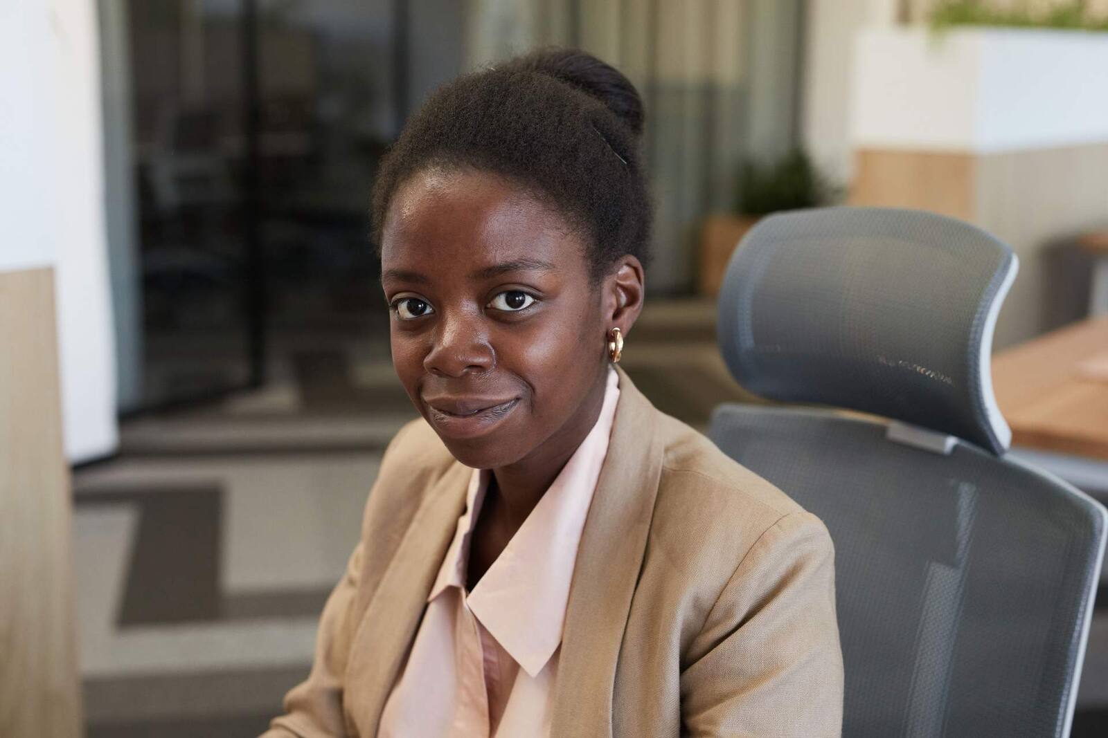A businesswoman in a tan blazer sits in an office chair, looking toward the camera
