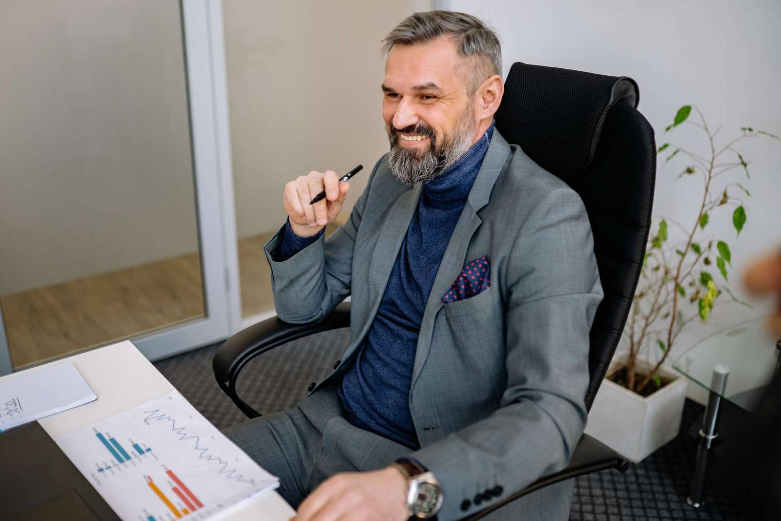 A gray-haired executive in a blazer smiles while reviewing bar and line charts on a printed report at a glass-topped desk