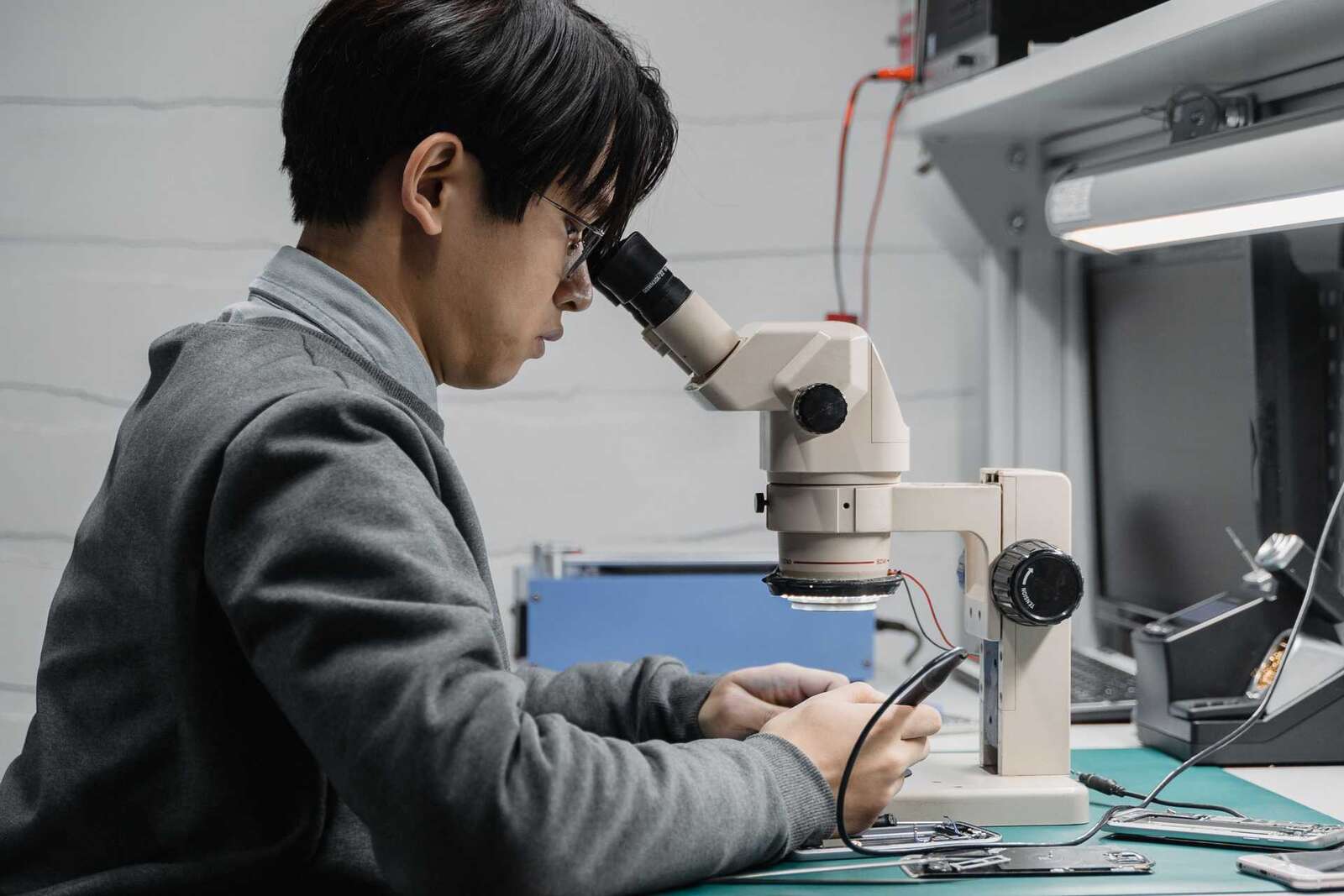 A technician in a gray sweater leans over a stereo microscope at a lab bench, examining a circuit board under bright overhead light