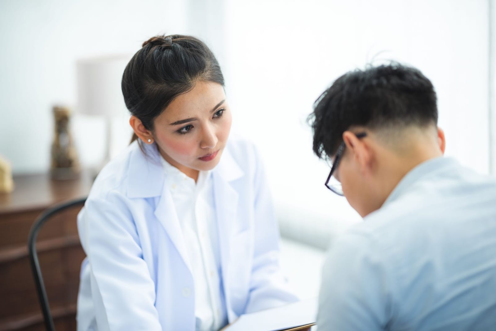 A physician in a white coat listens closely during a consultation with a patient in a bright exam room