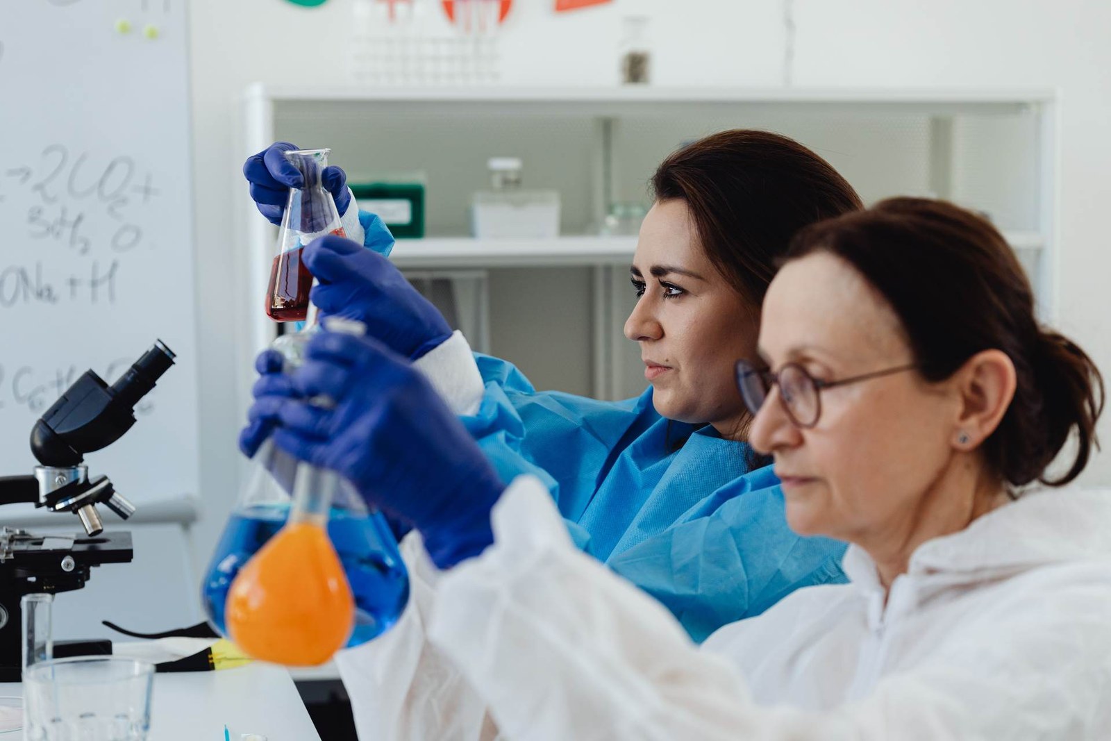 Two researchers in white and blue lab coats examine chemical flasks on a laboratory bench