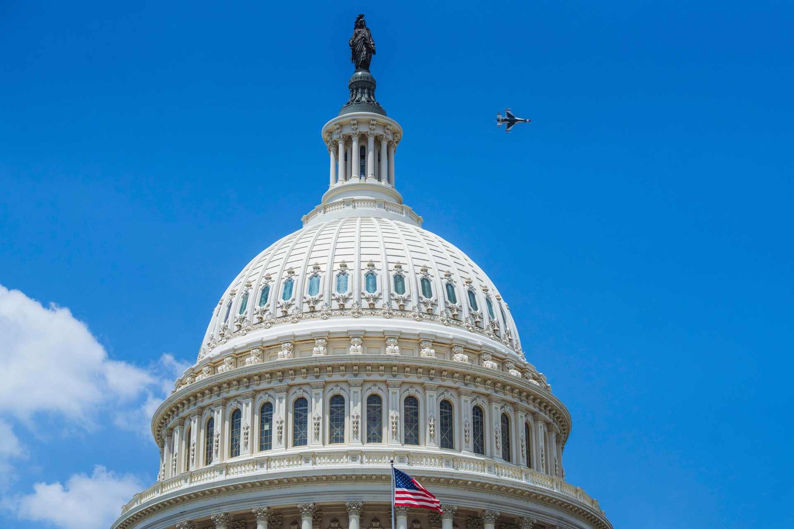 The dome of the United States Capitol against a clear blue sky, with a jet aircraft passing high to its right and a flag below