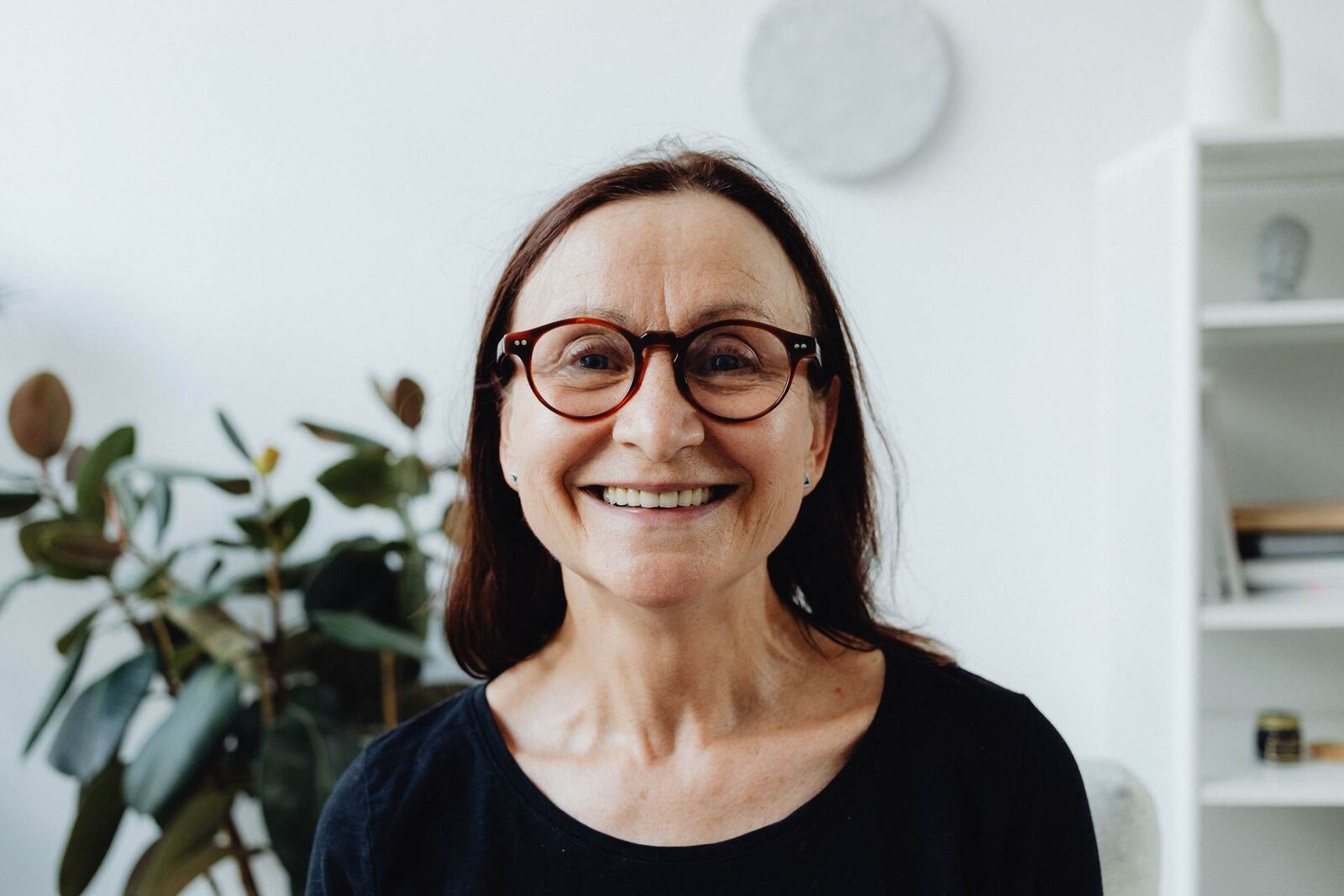 A smiling woman wearing glasses in a bright home living room, a generic stand-in for a patient managing care at home