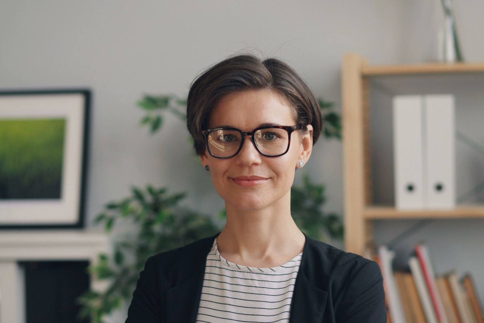 A professional wearing glasses and a dark blazer in an office with bookshelves and plants in the background