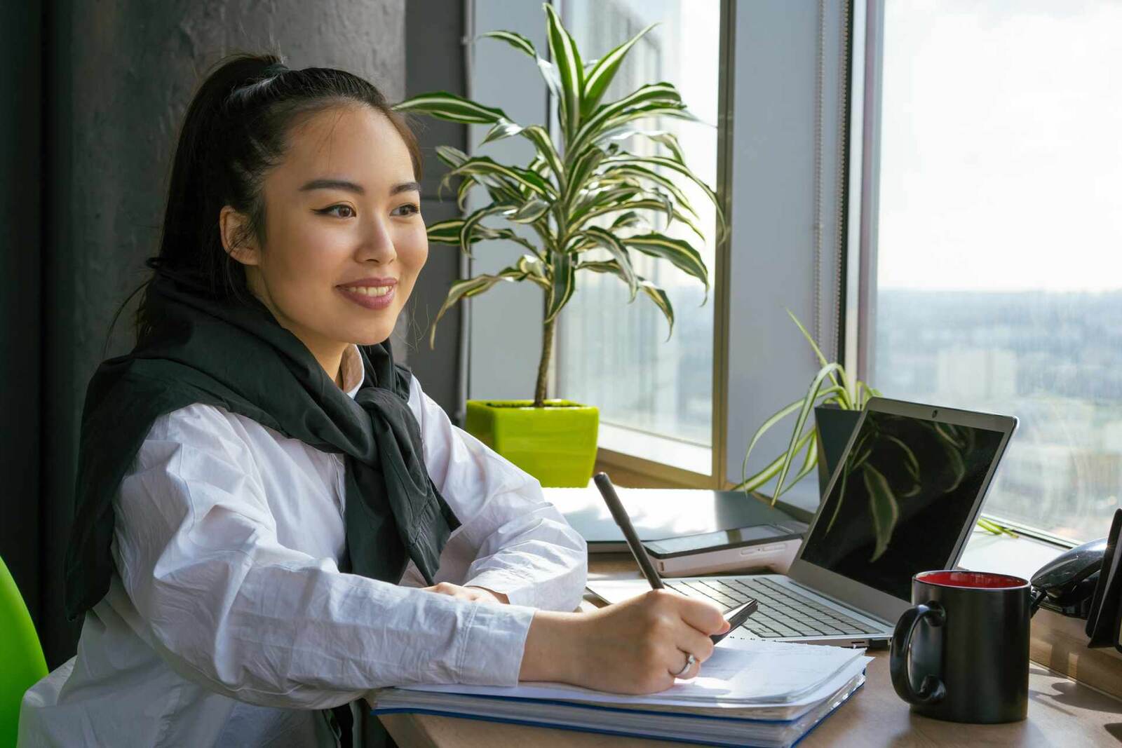 A professional reviews documents and a laptop at a bright office desk by a window