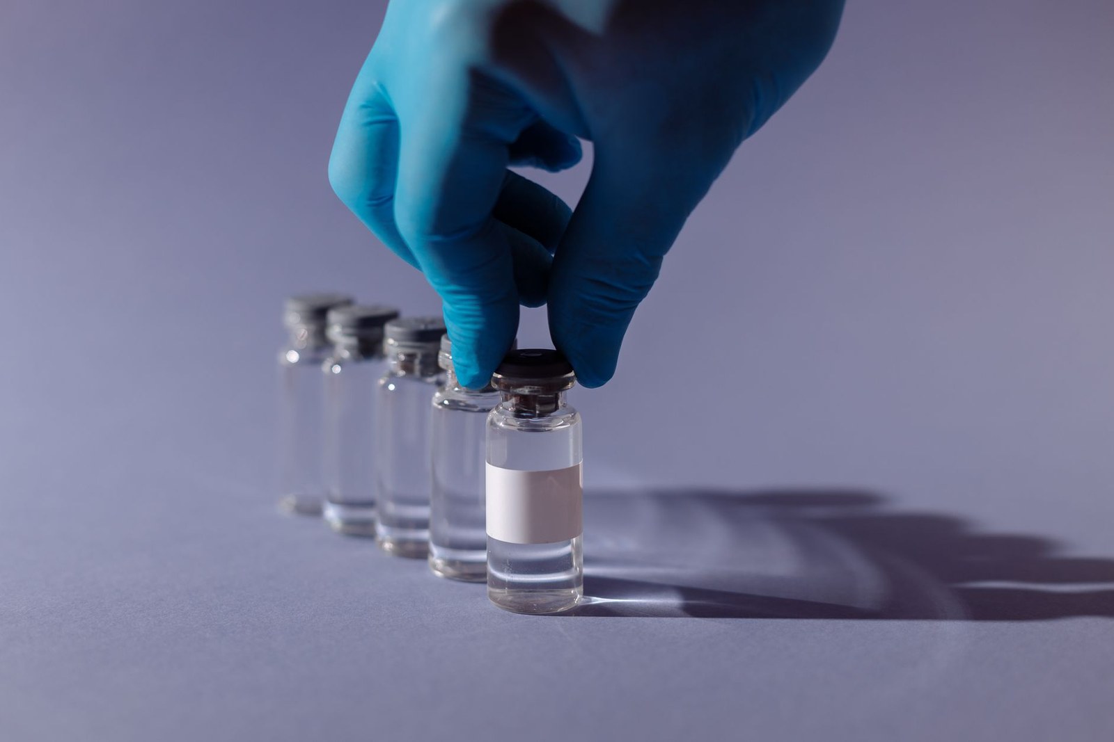 A gloved hand lifts a glass vial from a row of vials on a laboratory bench, its label left blank