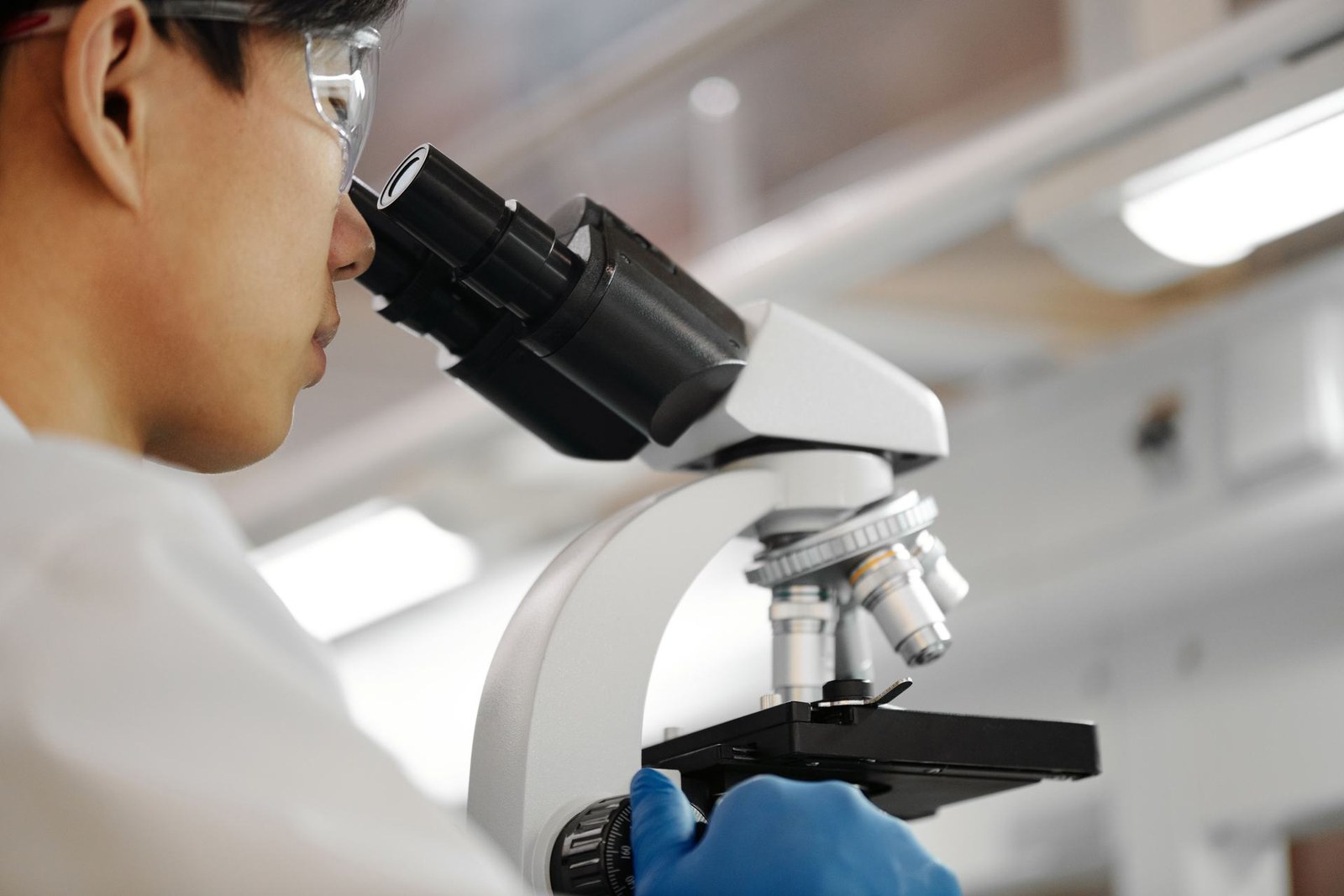 A researcher wearing safety glasses and a white lab coat looks through a microscope eyepiece while adjusting a sample slide