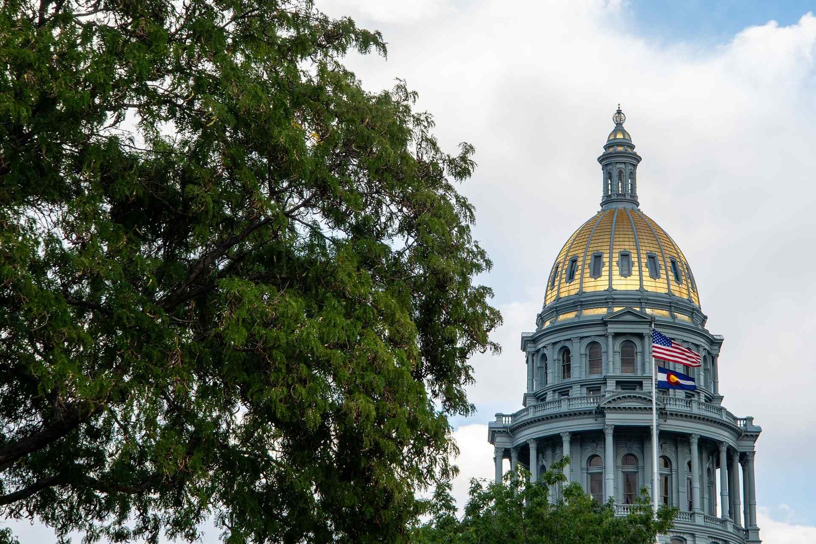 A gold-domed capitol building with an American flag rises above green trees against a partly cloudy sky