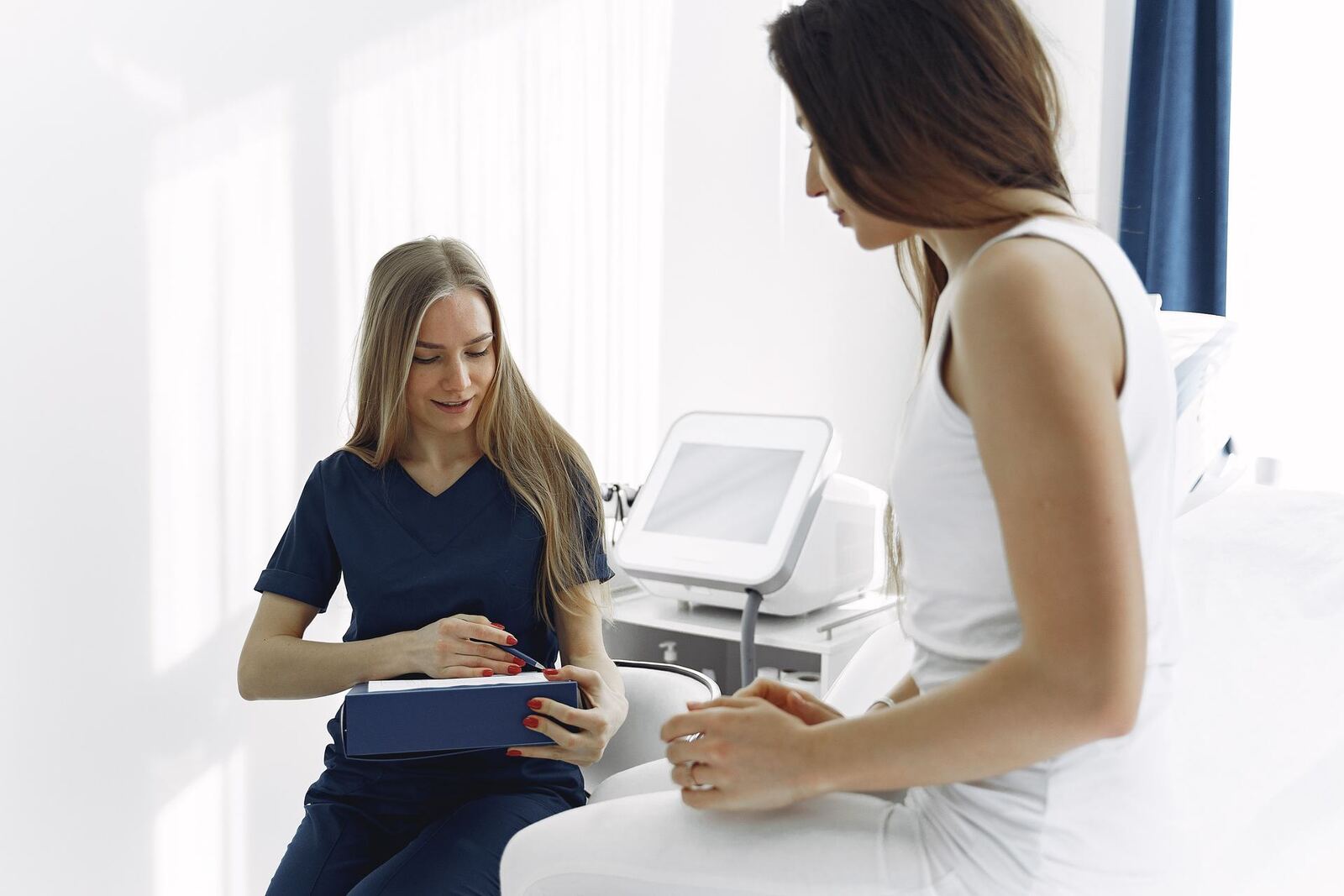 A clinician in scrubs consults with a patient beside a bedside monitor