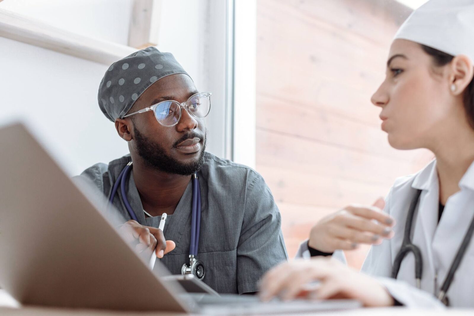Two clinicians in scrubs and a lab coat discussing a case over a laptop by a bright window, a generic stand-in for clinical and regulatory coordination