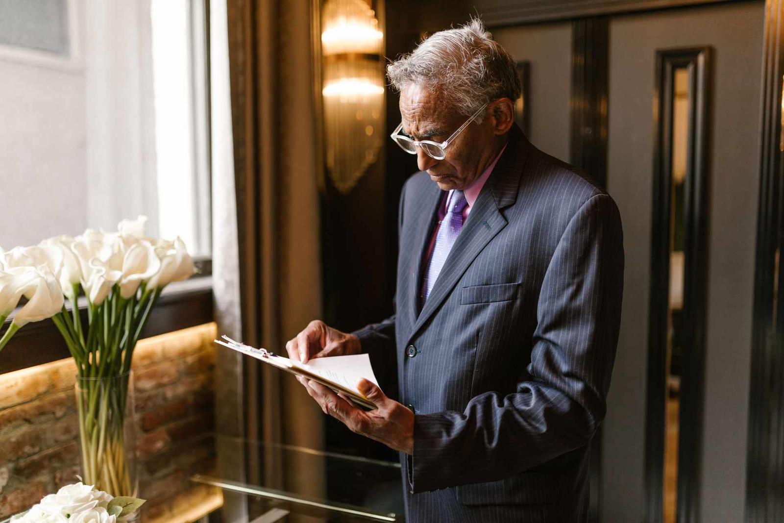 A senior professional in a pinstripe suit reads a clipboard document closely in a sunlit building lobby