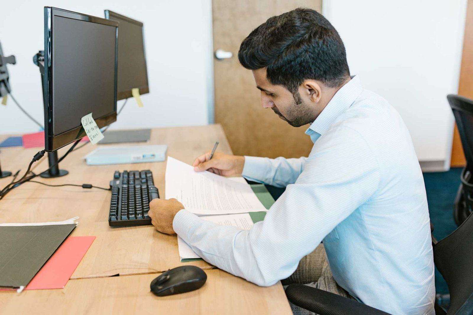 A man at a desk with two monitors annotates a printed document with a pen, folders and a mouse nearby