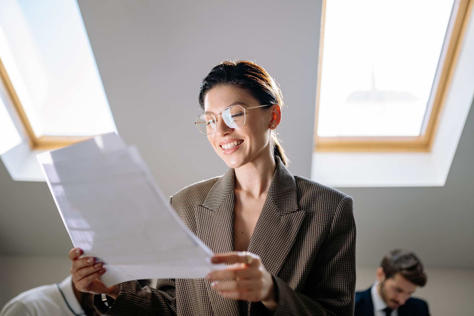 A smiling professional in glasses and a tweed blazer reads a printed document by a sunlit skylight window