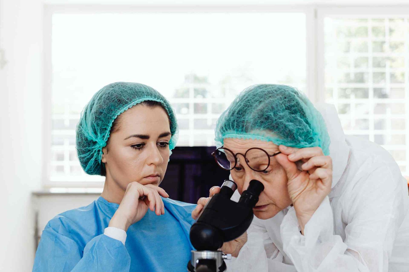 Two gowned, hairnet-capped laboratory researchers examining a sample under a microscope, one adjusting her glasses to look into the eyepiece